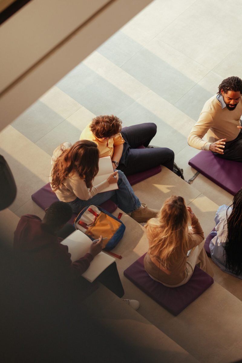 Group of diverse university students taking notes while listening to their teacher explaining a lesson, sitting on cushions in a modern building